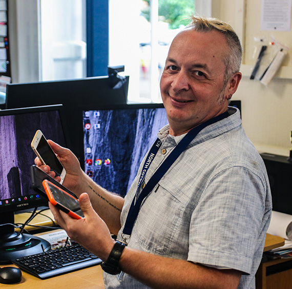 Photo of our link worker smiling and holding two mobile phones sitting at a desk with lots of computer monitors