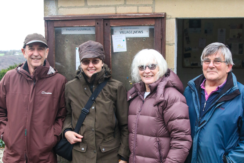 Four people standing in front of a village noticeboard which is attached to a bus shelter in Frampton Mansell