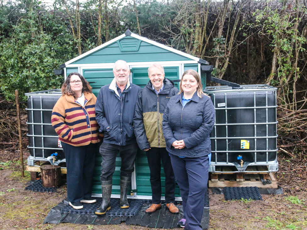 Parish Council and GRCC representatives standing in front of a green shed which has two very large water tanks to collect rainwater from its roof, in the village's community orchard